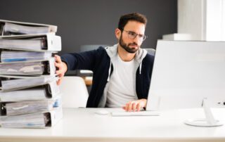 A man sitting at a desk using management software to organize a stack of folders in front of him.